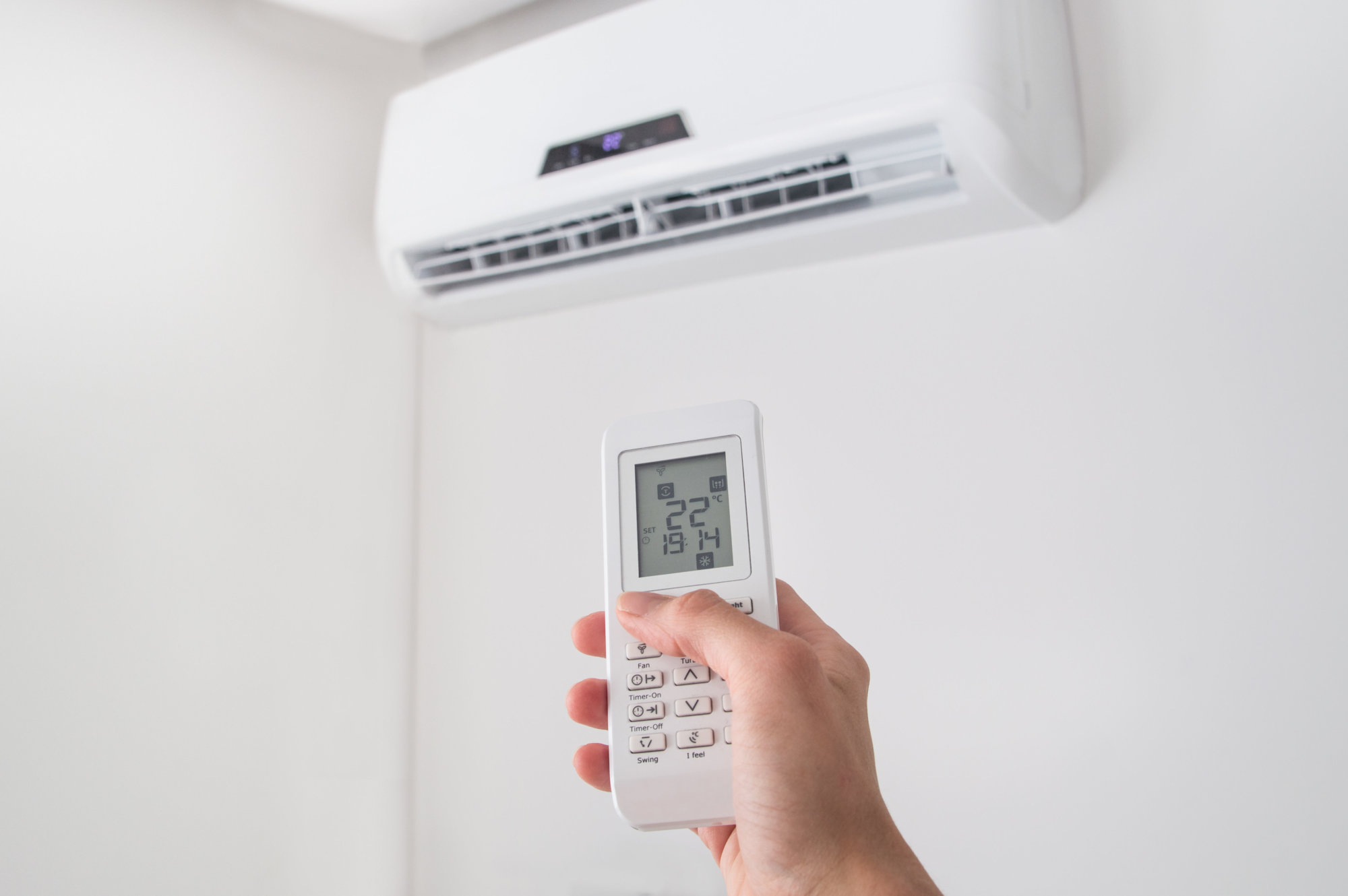 Hand holding remote control for air conditioner on white wall.