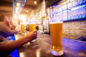 Close up view of hand while holding draft beer glass placed on a bar table with a row of other in the background.