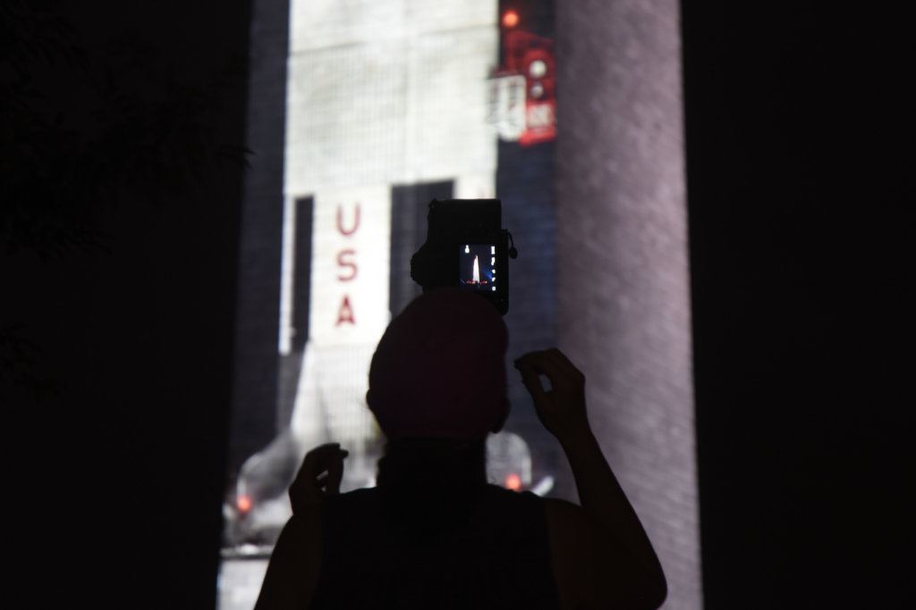 A man takes a photo of the projection of the rocket on the Washington Monument.