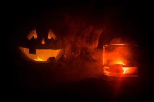 Halloween pumpkin with carved face and glass of whiskey with ice on a dark toned foggy background with zombies. Decorated. Selective focus