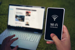 Smartphone with wifi connection generic icon on the screen hold by man on the background of laptop display and keyboard. Man uses Wi Fi hot spot to get to domestic internet access. Wireless gadgets