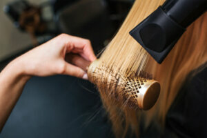 Female client in beauty salon. Close up of hairdressers hand drying blond hair with hair dryer and round brush, doing new hairstyle