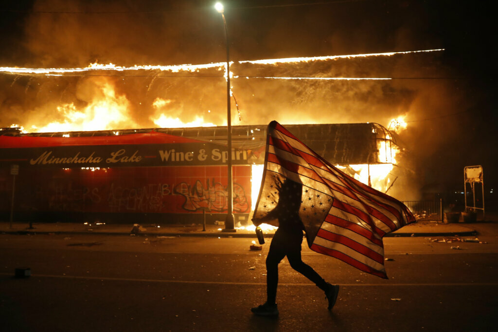 A protester carries the carries a U.S. flag upside, a sign of distress, next to a burning building Thursday, May 28, 2020, in Minneapolis. Protests over the death of George Floyd, a black man who died in police custody Monday, broke out in Minneapolis for a third straight night. (AP Photo/Julio Cortez)