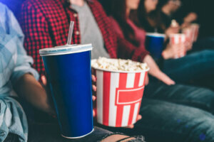 Close up of tasty but unhealthy food. There are basket of popcorn and a blue cup of coke on picture. Man and woman are holding it together.