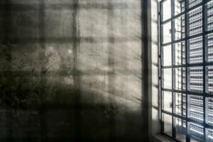 The very sober interior of a prison cell: barred windows with little light coming in and bare concrete walls