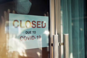 Business office or store shop is closed/bankrupt business due to the effect of novel Coronavirus (COVID 19) pandemic. Unidentified person wearing mask hanging closed sign in background on front door.