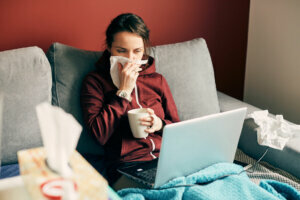 Sick girl works remotely at home with cup of tea and tissues on the bed