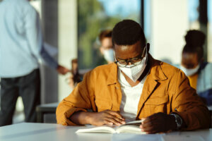 African American college student with face mask reading a book in the classroom.
