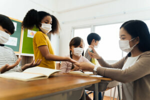 Young female teacher using an alcohol spray to disinfect student hands in classroom. Asian woman in face mask cleaning pupils' hands with hand sanitizer. School reopen after quarantine and lockdown