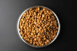 Dry pet food in a metal bowl on black background, top view