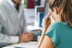 Woman suffering from bad headache or migraine. Appointment with doctor in office room. Sick and unwell lady with stress, trauma or burnout. Young patient in pain holding head with hands.