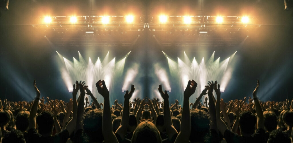 Concert stage, people are visible waving and clapping, silhouettes are visible
