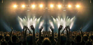 Concert stage, people are visible waving and clapping, silhouettes are visible