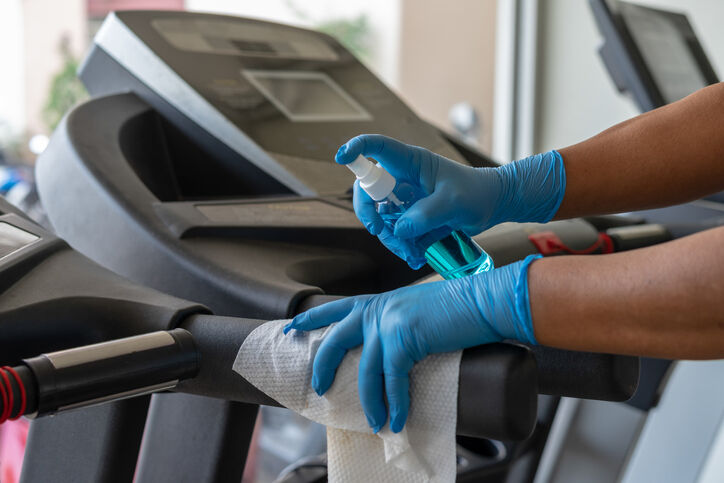 Staff using wet wipe and a blue sanitizer from the bottle to clean treadmill in gym.
