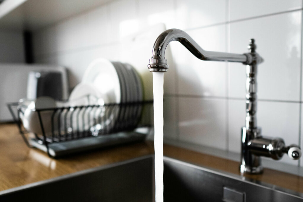 Water flowing out of a kitchen stainless steel tap into the sink. Wasting water by leaving a chrome faucet tap running. Water misuse in domestic duties and activities. Overusing household water.