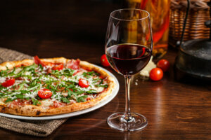Italian food. Classic thin pizza with large sides, prosciutto, cherry tomatoes, arugula, parmesan cheese. Serving dishes in a restaurant on a white plate with red wine.