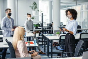 Corporate meeting and group work in company. African american woman manager in protective mask holding tablet, talking to workers keeping social distance