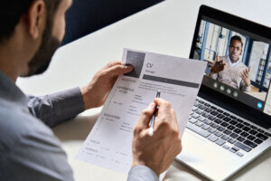Employer holding CV having online virtual job interview meeting with candidate on video call. Distance remote recruitment conference chat. Over shoulder view.