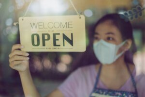 Close up shot of woman wearing mask and hand turning open sign board on glass door in coffee shop and restaurant after coronavirus lockdown quarantine.Business crisis concept.