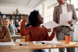 Happy African American student celebrating while receiving exam results from her professor in he classroom.