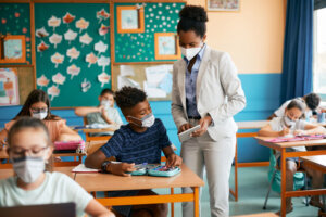 African American teacher and her student using touchpad and wearing protective face masks due to coronavirus pandemic.
