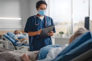 Hospital Ward: Friendly Nurse Wearing Face Mask Talks to Beautiful Senior Female Patient Resting in Bed, Checks Her Vitals, Writes down Information. Head Nurse and Old Lady Recovering after Covid