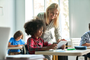 Female teacher helping African school girl using tablet at class in classroom.
