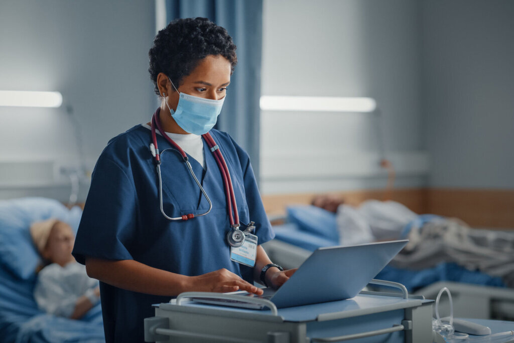 Hospital Ward: Professional Black Latin Female Head Nurse or Doctor Wearing Face Mask Uses Medical Computer. In the Background Patients in Beds Recovering Successfully after Sickness and Surgery