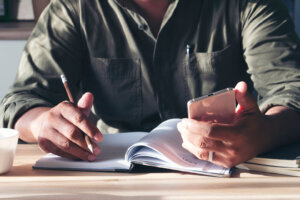Male hand of planner writing daily appointment On Calendar book, working space at home. Man plan and noted schedule (holiday trip) on diary and using smartphone at office desk. Planner concept.
