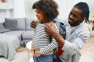 Happy daddy helps daughter getting ready for school and puts a school bag on her back