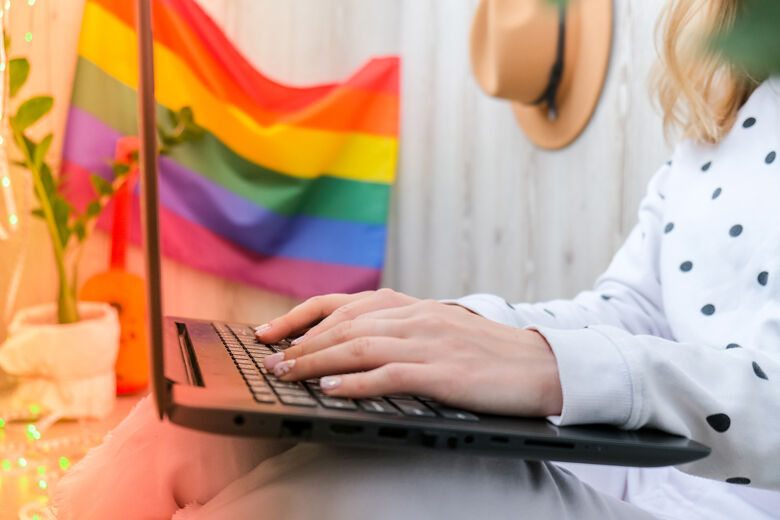 Young millennial hippie woman sitting on balcony using laptop. LGBTQ rainbow flag on background. Online education tuition. Work from home. Modern office