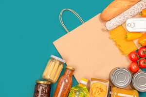 Food donations and paper bag on green background with copy space pasta, fresh vegatables, canned food, baguette and other groceries. Food bank, food delivery concept. Mockup
