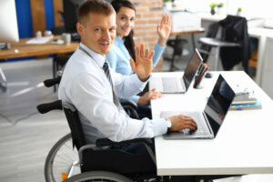 Businessman in wheelchair and businesswoman at table are smiling and raised their hands in greeting