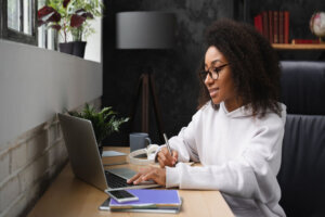 African young teenager e learning on distance, using laptop, writing homework assignment in copybook, watching webinars online from home.