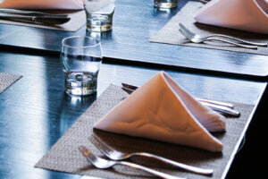 Folded white napkins on table mat, stainless steel knives forks and crystal drinking glasses on black wooden table
