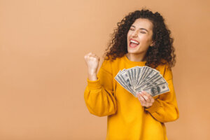 Portrait of a cheerful young woman holding money banknotes and celebrating isolated over beige background.