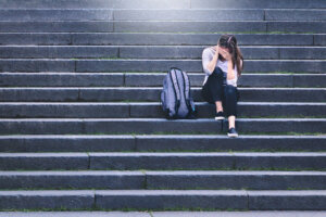 Bullying, discrimination or stress concept. Sad teenager crying in school yard. Upset young female student having anxiety. Upset victim of abuse or harassment sitting on stairs outdoors.