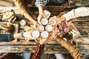 Friends cheering beer glasses on wooden table covered with delicious food Top view of people having dinner party at bar restaurant Food and beverage lifestyle concept