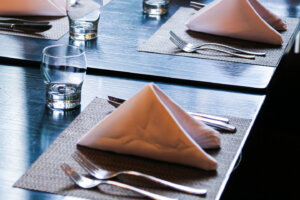 Folded white napkins on table mat, stainless steel knives forks and crystal drinking glasses on black wooden table