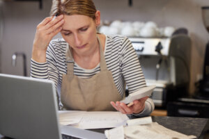 Worried Female Owner Of Coffee Shop In Financial Difficulty Looking Through Bills Using Laptop And Calculator