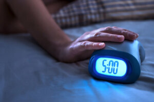 Tired woman waking up for work or school early in the morning. Grumpy lady pushing snooze button or turning off alarm clock with hand. Getting up after sleeping. Exhausted person in bed.