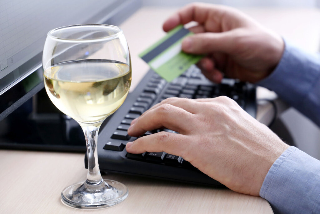 Man holding credit bank card types on PC keyboard, defocused view through glass of white wine