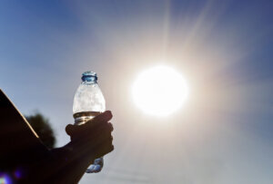 Bottle with water in a woman's hand against bright sun outdoors. Climate change hotness concept