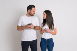 Serious boyfriend and girlfriend using mobile phone and looking at each other on white background