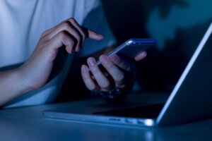 Close up young man hand holding smartphone for access by unlock on screen and using laptop for register website on internet in the late night , internet addiction concept