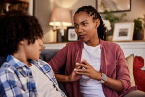 An African American woman sitting on a couch with her teenage son and talking.