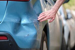 Driver hand examining dented car with damaged fender parked on city street side. Road safety and vehicle insurance concept