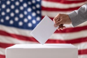 Election in USA. Man hand putting his vote into ballot box and American flag on background – Close up