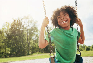 Happy, nature and child on swing in park for fun, adventure and play time on holiday or weekend trip. Smile, cute and boy kid on motion equipment on playground in outdoor field on vacation