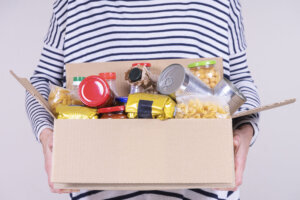 Volunteer hands holding food donations box with grocery products on white desk